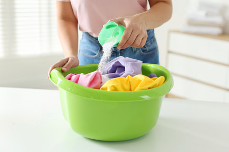 Woman pouring washing powder into plastic basin with clothes at white table indoors, closeup