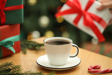 Woman holding gift box and wooden table with aromatic coffee, candy cane and fir tree branches against blurred lights, selective focus. Christmas greeting cardの写真素材