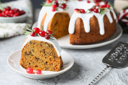 Piece of delicious Christmas cake with icing, cranberries and rosemary on light gray textured table,...