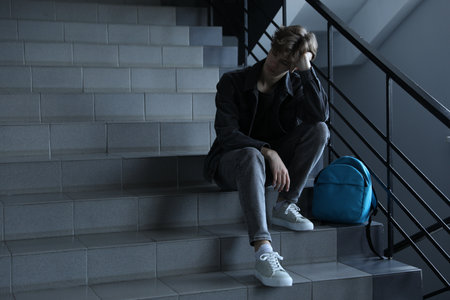 Depressed teenage boy and backpack on stairs indoors, space for textの写真素材