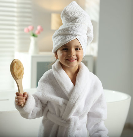 Cute girl in bathrobe with wrapped hair and brush near bathtub indoorsの写真素材