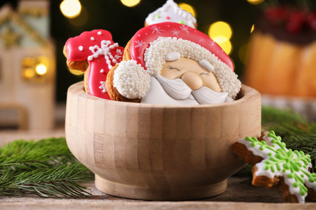 Tasty gingerbread cookies in bowl and fir tree branches on wooden table against blurred lights, closeup. Christmas treatの写真素材