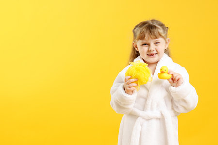 Happy girl in bathrobe with loofah and rubber duck on yellow background. Space for textの写真素材