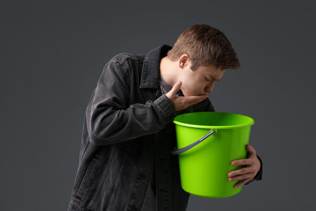 Young man with bucket suffering from nausea on gray backgroundの写真素材
