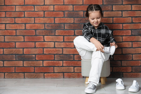 Little girl trying on different shoes near brick wall indoors. Space for textの写真素材
