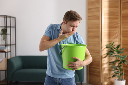 Young man with bucket suffering from nausea indoorsの写真素材