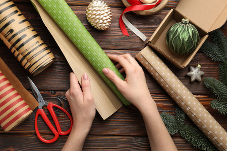 Festive packaging. Woman with gift box, ribbon, scissors, wrapping paper and Christmas decor at wooden table, top viewの写真素材