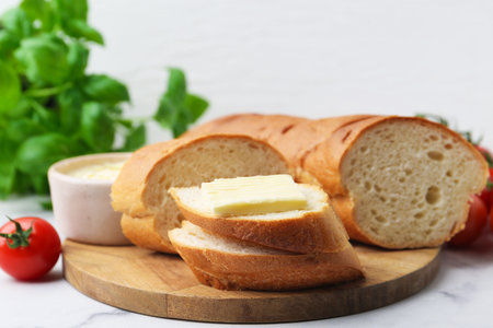 Cut baguette with butter, basil and tomato on white marble table, closeup