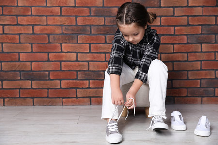 Little girl trying on different shoes near brick wall indoors. Space for textの写真素材