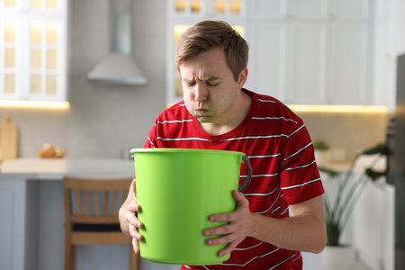 Young man with bucket suffering from nausea indoorsの写真素材