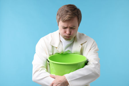 Young man with bucket suffering from nausea on light blue backgroundの写真素材