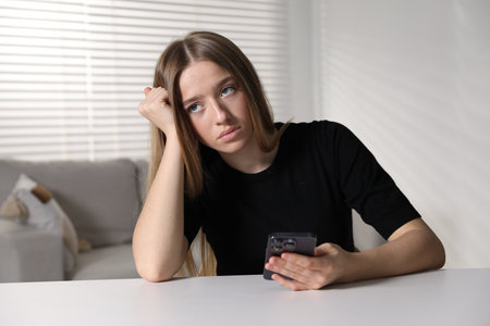 Sad teenage girl with smartphone at white table indoorsの写真素材