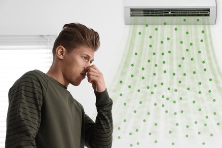 Young man suffering from smell from air conditioner and covering his nose indoors. Air flows with bacteria from applianceの写真素材