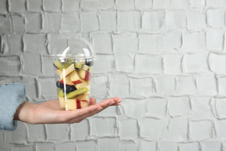 Woman holding plastic cup of fresh fruit salad against light brick wall, closeup. Space for textの写真素材