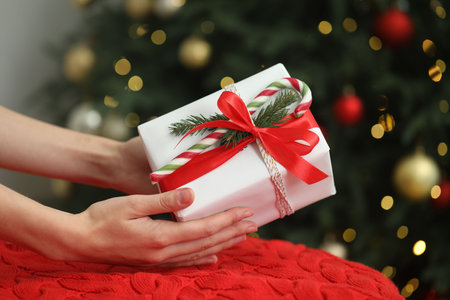 Woman holding decorated gift with candy cane near Christmas tree, closeup