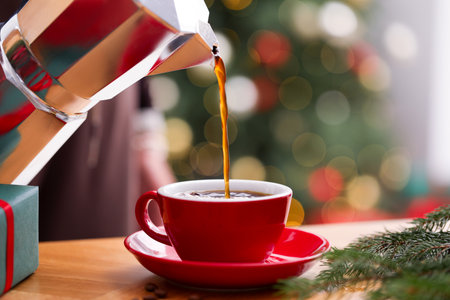 Pouring aromatic coffee from moka pot into cup at wooden table with gift box and fir tree branches against blurred lights, closeup. Christmas greeting cardの写真素材