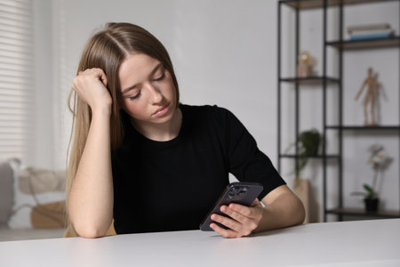 Sad teenage girl with smartphone at white table indoorsの写真素材