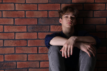 Depressed teenage boy near brick wall at home, space for text