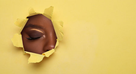 Young woman with bright makeup, closeup. View on eye through torn hole in yellow paperの写真素材