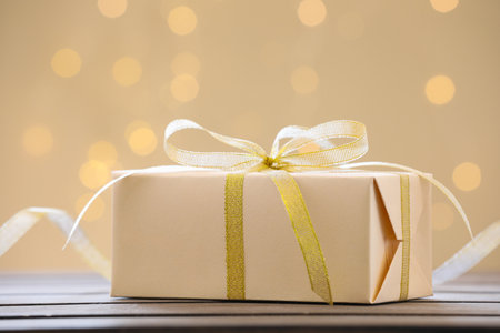 Beautiful gift box with ribbon on wooden table against golden background with blurred lights,...