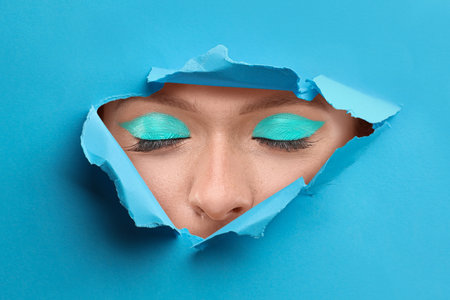 Young woman with bright makeup, closeup. View on eyes through torn hole in blue paperの写真素材