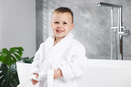 Smiling little boy in white bathrobe leaning on bathtub in bathroomの写真素材