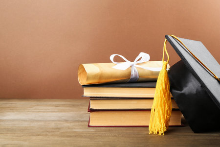 Graduation cap, diploma and books on wooden table against brown background, closeup. Space for textの写真素材
