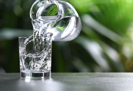 Pouring water into glass from jug at gray table against blurred green background, closeup. Space for...