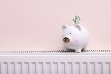 Piggy bank and banknote on heating radiator near beige wall, closeup. Space for textの写真素材