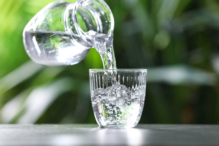 Pouring water into glass from jug at gray table against blurred green background, closeup. Space for textの写真素材