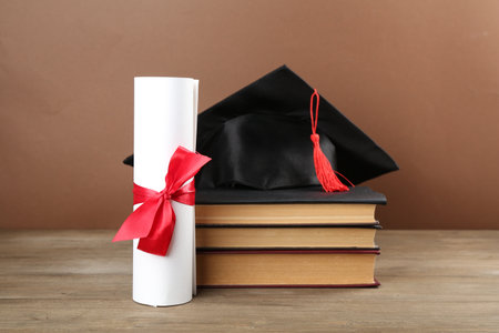 Graduation cap, diploma and books on wooden table against brown backgroundの写真素材