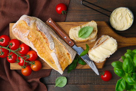 Slices of baguette with butter, basil, tomatoes and knife on wooden table, flat layの写真素材