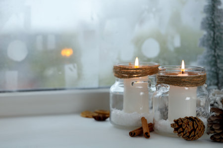 DIY Christmas lanterns. Burning candles in glass jars, cones and spices on window sill indoors, closeup. Space for textの写真素材