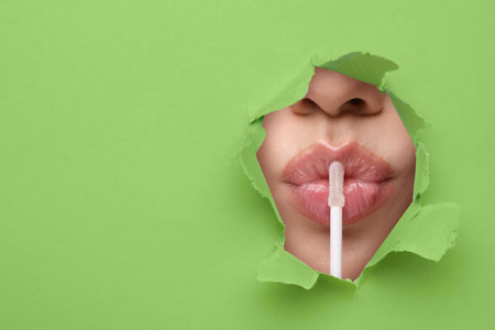 Young woman applying lip gloss, closeup. View on lips through torn hole in light green paperの写真素材