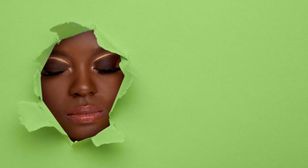 Young woman with stylish makeup, closeup. View on face through torn hole in light green paperの写真素材