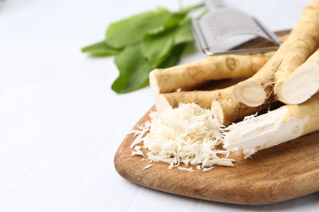Grated horseradish, grater, leaves and fresh roots on white tiled table, closeup. Space for textの写真素材