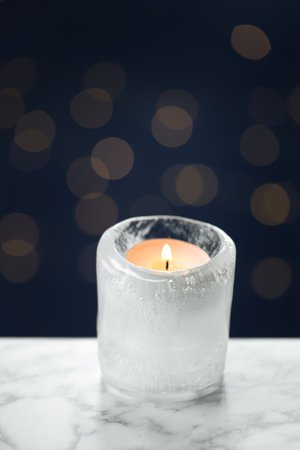 DIY Christmas lantern. Burning ice candle on white marble table against blue background with blurred lights, closeup. Space for textの写真素材