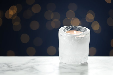 DIY Christmas lantern. Burning ice candle on white marble table against blue background with blurred lights, closeup. Space for textの写真素材