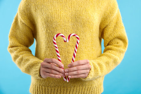 Woman in yellow sweater heart making with candy canes on light blue background, closeup. Merry Christmas and Happy New Yearの写真素材