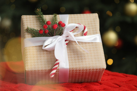 Decorated Christmas gift with candy cane on plaid against blurred lights, closeup
