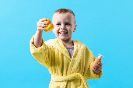 Smiling little boy in yellow bathrobe with rubber duck and bottle of cosmetic product on light blue backgroundの写真素材
