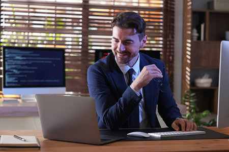 Smiling programmer in suit with glasses working on laptop at wooden table in office