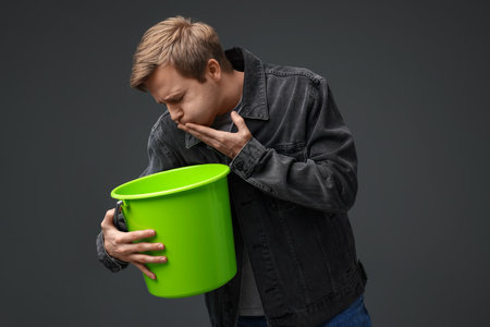 Young man with bucket suffering from nausea on gray backgroundの写真素材