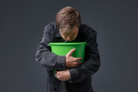 Young man with bucket suffering from nausea on gray backgroundの写真素材