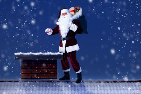 Santa Claus with bag full of gifts near chimney on roof of house at nightの写真素材