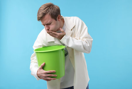 Young man with bucket suffering from nausea on light blue backgroundの写真素材