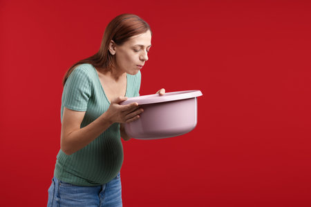 Pregnant woman with basin suffering from nausea on red background. Space for textの写真素材