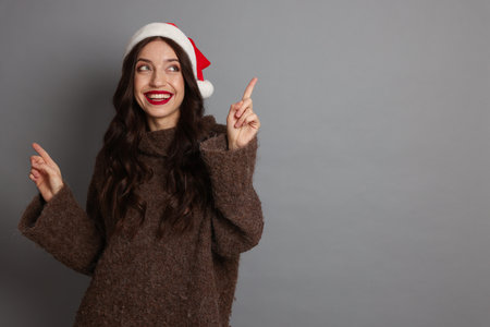 Beautiful woman with Santa hat pointing at something on gray background, space for text. Christmas seasonの写真素材