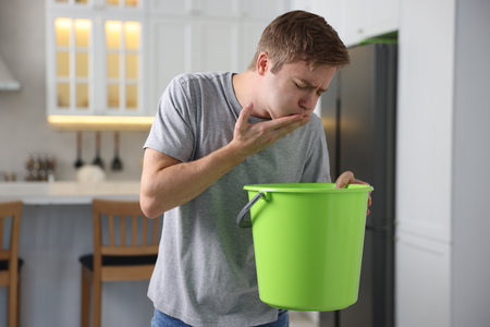 Young man with bucket suffering from nausea indoorsの写真素材