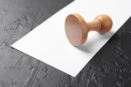 One wooden stamp and blank sheet of paper on dark textured table, closeup. Space for textの写真素材